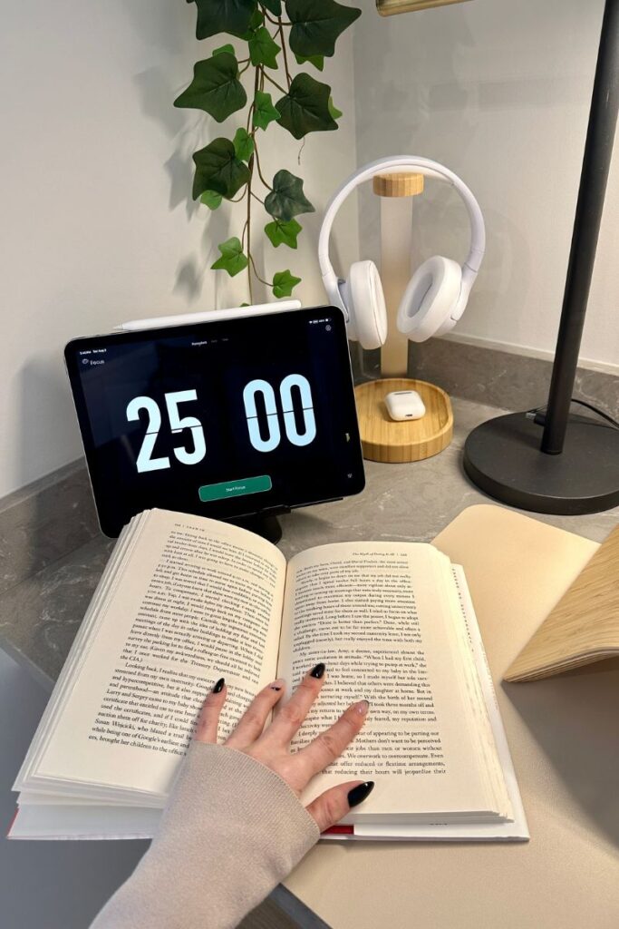 A female hand holding open a book in from of an iPad with 25:00 displayed. Plant and headphones in the background.