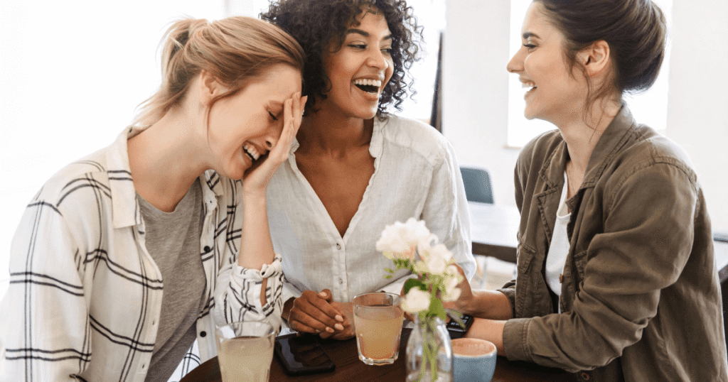 Women laughing around a table at a cafe with drinks on table. Credit @dean-drobot
