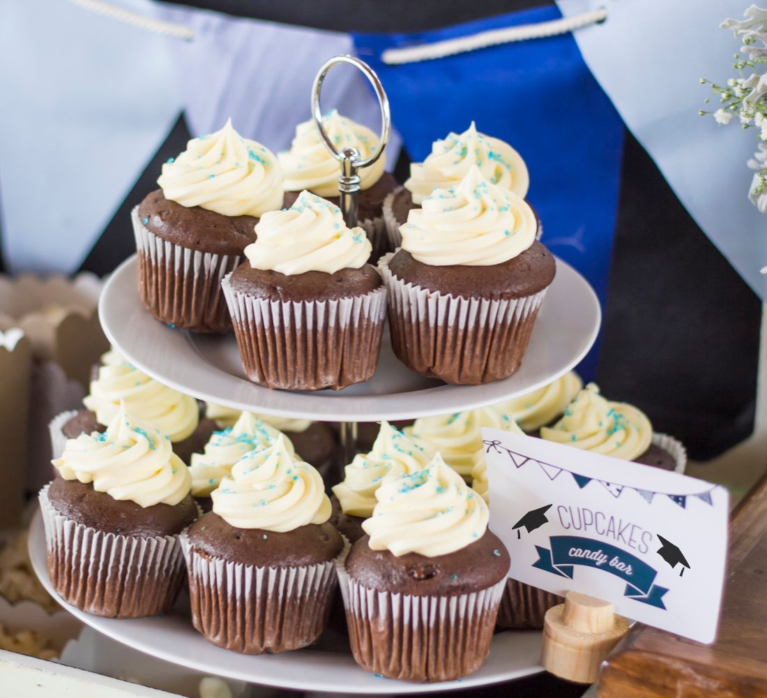 Chocolate cupcakes with white icing and blue sprinkles on a 2-tier rack with a sign that reads "Cupcakes - Candy Bar"