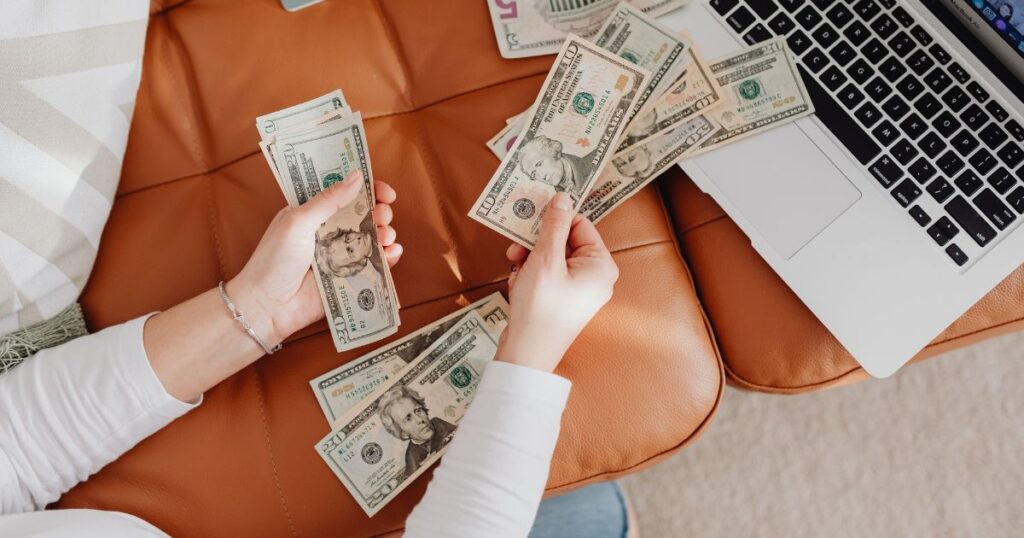 A woman with handfuls of cash sitting on her couch with an open laptop