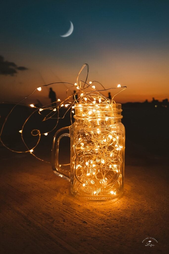A mason jar centerpiece filled with fairy lights on an outdoor table with a clear moon sky in the background.